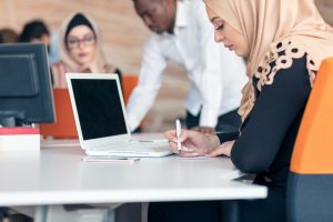 a young arabic woman with a hijab is writing at a table, her laptop is open but not on. A group of people work around her. SEO trends for 2025 are important to watch and adjust to.